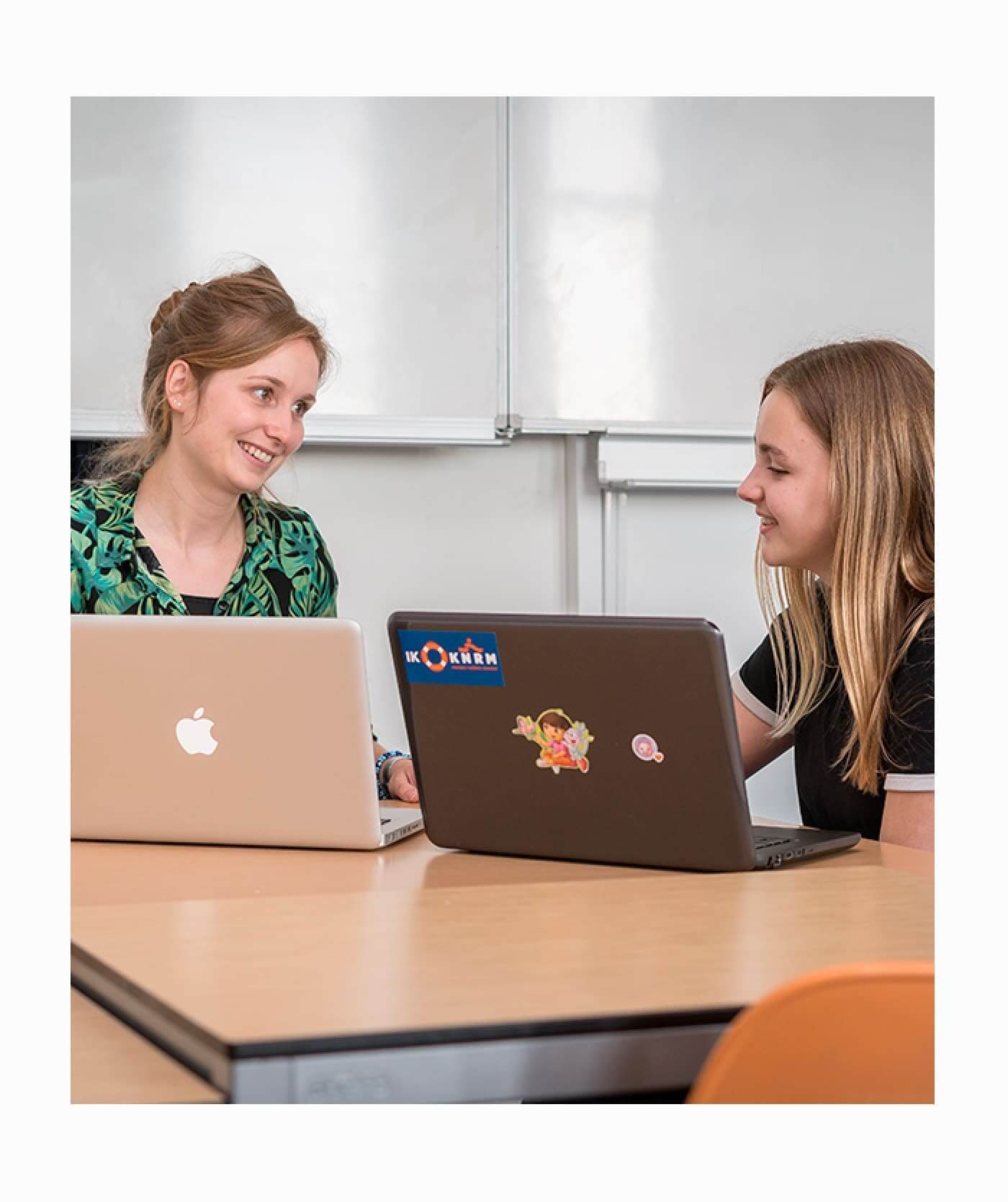 Twee vrouwen zitten in een klaslokaal met een whiteboard aan een tafel met laptops en lachen naar elkaar. In een vriendelijke, professionele sfeer werken ze samen aan school.
