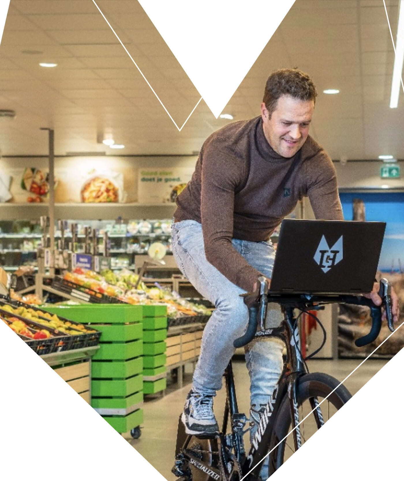 Een man fietst binnenshuis op een hometrainer en gebruikt een laptop op het stuur om websites te bekijken. Hij staat in de groente- en fruitafdeling van een supermarkt.