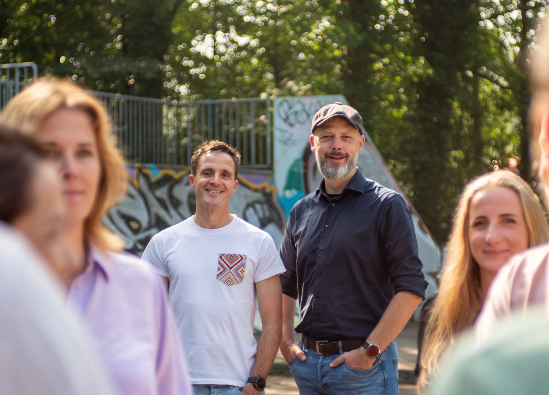 Verschillende volwassenen staan buiten voor met graffiti bedekte bouwwerken en bomen. Twee mannen zijn scherp op de voorgrond en anderen zijn vaag op de foto te zien. Het doet denken aan creatieve teams achter innovatieve websites of online diensten.