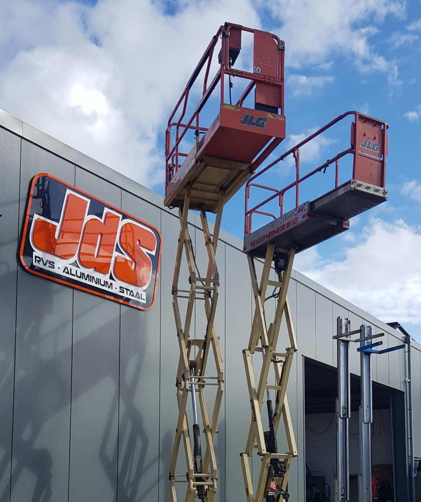 Naast een bedrijfspand staan twee schaarliften met een JdS RVS·ALUMINIUM·STAAL bord. Onder een deels bewolkte hemel benadrukken zij hun expertise op het gebied van webshops en online diensten.